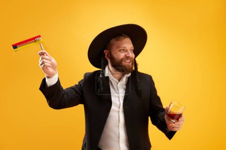 Téléchargez les photos : Joyeux jeune homme juif au chapeau, avec des latéraux tenant bruyant, sur fond jaune. Fête. Concept de vacances Pourim, traditions juives, histoire et culture - en image libre de droit