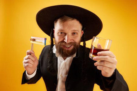 Téléchargez les photos : Heureux homme juif barbu en chapeau avec sidelock, tenant bruyant et verre de cognac, célébrant sur fond de studio jaune. Concept de vacances Pourim, traditions juives, histoire et culture - en image libre de droit