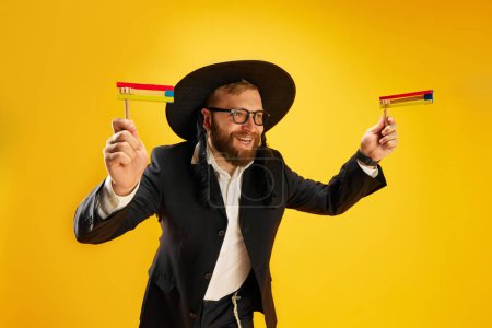 Photo for Bearded young Jewish man in glasses, yarmulke playing with wooden noisemaker, gragger against yellow studio background. Concept of Purim holiday, Jewish traditions, history and culture - Royalty Free Image