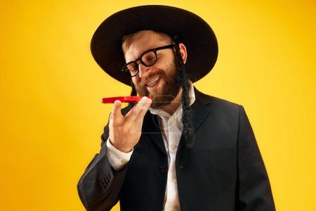 Photo for Bearded young man in hat with sidelock recording voice message on phone, greeting with holiday against yellow studio background. Concept of Purim holiday, Jewish traditions, history and culture - Royalty Free Image