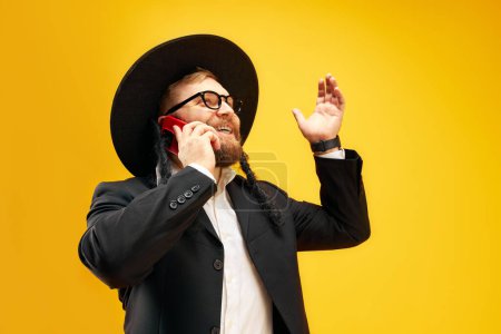 Photo for Bearded young man in hat with sidelock emotionally talking on phone, greeting with holiday against yellow studio background. Concept of Purim holiday, Jewish traditions, history and culture - Royalty Free Image