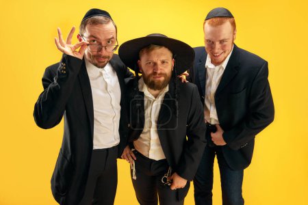 Téléchargez les photos : Trois hommes juifs, amis aux attributs juifs traditionnels souriants, posant sur fond de studio jaune. Concept de vacances Pourim, traditions juives, histoire et culture - en image libre de droit