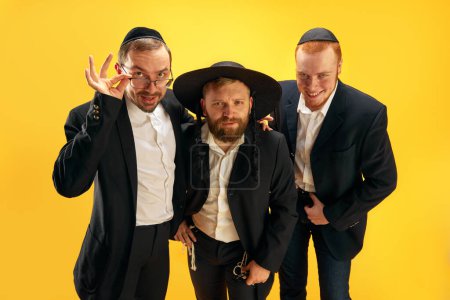 Photo for Three Jewish men, friends in traditional Jewish attributes smiling, posing against yellow studio background. Concept of Purim holiday, Jewish traditions, history and culture - Royalty Free Image