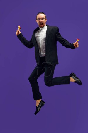 Photo for Full-length of emotional Jewish man in his 30s wearing black suit, holding noisemaker and jumping against purple studio background. Concept of Purim holiday, Jewish traditions, history and culture - Royalty Free Image