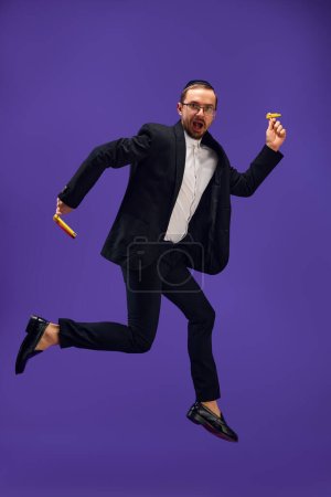 Photo for Full-length of emotional Jewish man in his 30s wearing black suit, holding noisemaker and jumping against purple studio background. Concept of Purim holiday, Jewish traditions, history and culture - Royalty Free Image