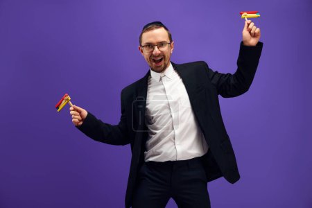 Photo for Positive, excited young Jewish man in in glasses standing with noisemaker against purple studio background. Happy. Concept of Purim holiday, Jewish traditions, history and culture - Royalty Free Image