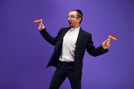 Photo for Positive, excited young Jewish man in in glasses standing with noisemaker against purple studio background. Happy. Concept of Purim holiday, Jewish traditions, history and culture - Royalty Free Image
