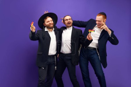 Photo for Enjoyment. Three Jewish men, friends in traditional Jewish attributes smiling, posing against against purple studio background. Concept of Purim holiday, Jewish traditions, history and culture - Royalty Free Image