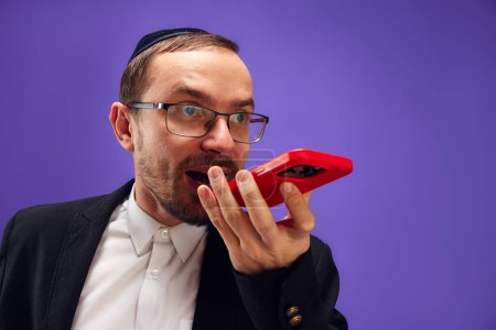 Photo for Emotional man in glasses and yarmulke recording voice message on phone, greeting with holiday against purple studio background. Concept of Purim holiday, Jewish traditions, history and culture - Royalty Free Image
