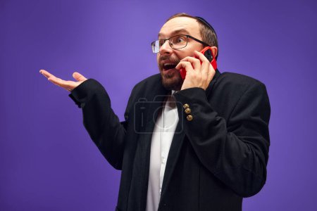 Photo for Excited man in glasses and yarmulke emotionally talking on phone, greeting with holiday against purple studio background. Concept of Purim holiday, Jewish traditions, history and culture - Royalty Free Image