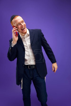 Photo for Young redhead emotional Jewish man emotionally talking on phone, greeting with holiday against purple studio background. Concept of Purim holiday, Jewish traditions, history and culture - Royalty Free Image