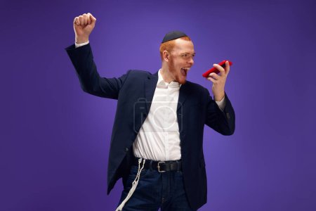 Photo for Young Jewish smiling man emotionally recording voice message against purple studio background. Concept of Purim holiday, Jewish traditions, history and culture - Royalty Free Image