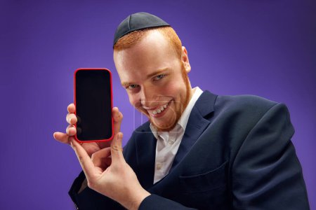 Téléchargez les photos : Jeune juive émotionnelle ma montrant écran de téléphone portable sur fond violet studio. Des nouvelles. Concept de vacances Pourim, traditions juives, histoire et culture - en image libre de droit