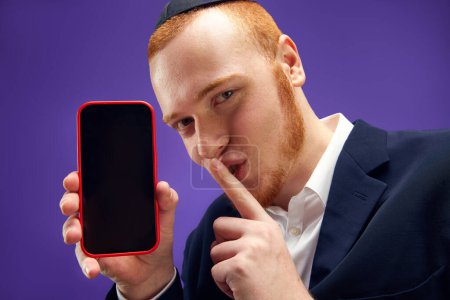 Photo for Young Jewish emotional ma showing mobile phone screen against purple studio background. News. Concept of Purim holiday, Jewish traditions, history and culture - Royalty Free Image