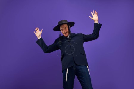 Téléchargez les photos : Joyeux et excité jeune homme juif avec des sidelocks dans le chapeau, costume posant sur fond violet studio. Concept de vacances Pourim, traditions juives, histoire et culture - en image libre de droit