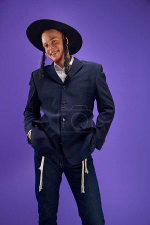 Téléchargez les photos : Joyeux et excité jeune homme juif avec des sidelocks dans le chapeau, costume posant sur fond violet studio. Concept de vacances Pourim, traditions juives, histoire et culture - en image libre de droit