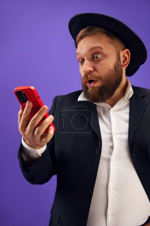 Photo for Bearded young Jewish man emotionally looking on mobile phone screen, reading news and messages against purple studio background. Concept of Purim holiday, Jewish traditions, history and culture - Royalty Free Image