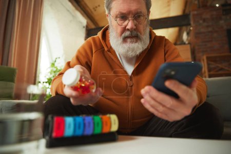 Foto de Hombre de edad avanzada examinando la botella de la píldora mientras se mira smartphone. Usando organizador de pastillas. Servicios médicos en línea. Concepto de salud y atención médica, envejecimiento, medicina, tratamiento - Imagen libre de derechos