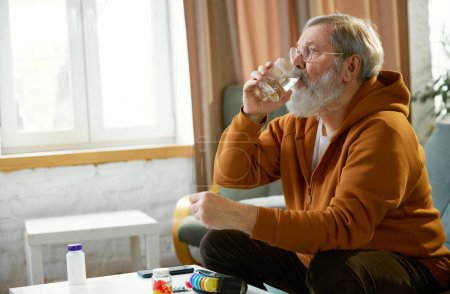 Téléchargez les photos : Homme âgé avec canne assis à côté de la table avec des médicaments, smartphone, et un verre d'eau. Prendre des médicaments. Concept de santé et de soins médicaux, vieillissement, médecine, traitement - en image libre de droit