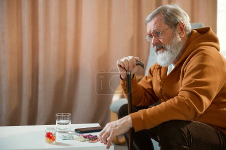 Photo for Senior man with cane sitting beside table with medications, smartphone, and a glass of water. Concentrated sad face. Meds. Concept of health and medical care, aging, medicine, treatment - Royalty Free Image