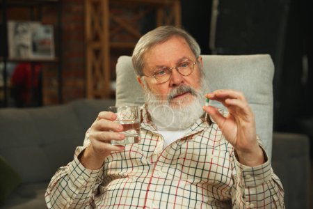 Téléchargez les photos : Homme âgé prenant la pilule avec un verre d'eau, assis confortablement dans un salon à la maison. Prendre soin de sa santé. Concept de santé et de soins médicaux, vieillissement, médecine, traitement - en image libre de droit