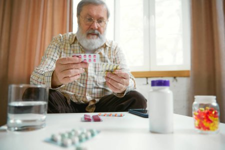Foto de Hombre mayor sentado en casa durante el día, sosteniendo pastillas, revisándolas. La importancia de cuidar altera la salud en la vejez. Concepto de salud y atención médica, envejecimiento, medicina, tratamiento - Imagen libre de derechos