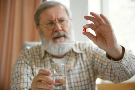 Téléchargez les photos : Homme âgé assis à la maison et buvant des pilules. Fond flou. Focus sur les mains avec capsule rouge, pilule. Des vitamines. Concept de santé et de soins médicaux, vieillissement, médecine, traitement - en image libre de droit