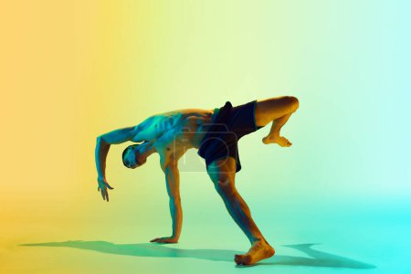 Foto de Una pose dinámica. Hombre joven musculoso y atlético entrenando sin camisa, practicando contra el degradado fondo amarillo azul en luz de neón. Concepto de estilo de vida activo y saludable, deporte, fitness, resistencia - Imagen libre de derechos