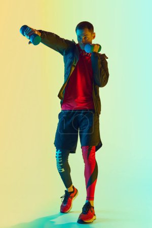 Téléchargez les photos : Exercices manuels. Homme athlétique en entraînement de vêtements de sport avec haltères contre fond bleu jaune dégradé au néon. Concept de mode de vie actif et sain, sport, fitness, endurance - en image libre de droit