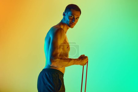 Photo for Athletic young man, with muscular, relief shirtless body training with resistance band against gradient blue yellow background in neon light. Concept of active and healthy lifestyle, sport, fitness - Royalty Free Image