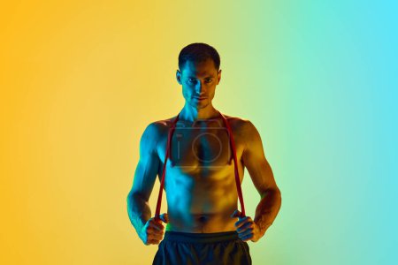 Photo for Handsome young man with muscular, shirtless, relief body posing wit fitness resistance band against gradient blue yellow background in neon light. Concept of active and healthy lifestyle, sport - Royalty Free Image
