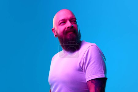 Téléchargez les photos : Portrait d'homme chauve barbu avec des tatouages en t-shirt blanc décontracté posant sur fond bleu au néon. Concept d'émotions humaines, expression faciale - en image libre de droit