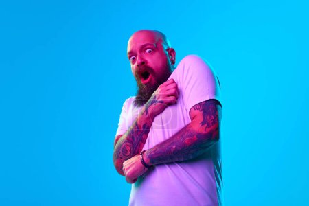 Photo for Portrait of emotional breaded bald man expressing fearful face, gesturing, shouting against blue background in neon light. Being afraid. Concept of human emotions, facial expression - Royalty Free Image