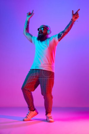 Foto de Imagen completa de hombre calvo barbudo en ropa casual, con tatuajes, escuchando música en auriculares contra el degradado fondo púrpura en luz de neón. Concepto de emociones humanas, expresión facial - Imagen libre de derechos