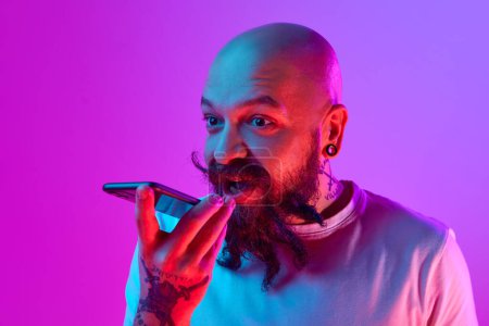 Photo for Bearded bald man with stylish moustaches recording voice message with phone against gradient purple background in neon. Concept of human emotions, facial expression, online convenient communication - Royalty Free Image