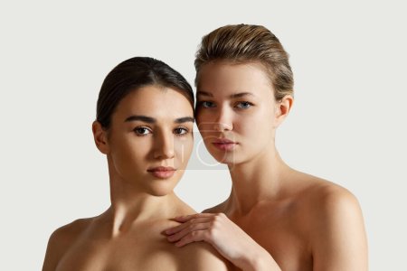 Photo for Beautiful two girls with different skin tones, perfect smooth spotless skin, with bare shoulders standing over white studio background. Concept of natural beauty, cosmetology and cosmetics, skincare - Royalty Free Image