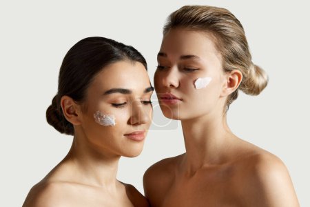 Photo for Blonde and brunette beautiful girls with spotless, clear, well-kept skin applying face moisturizing cream over white studio background. Concept of natural beauty, cosmetology and cosmetics, skincare - Royalty Free Image