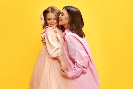 Photo for Portrait of beautiful young woman kissing her little pretty daughter, mother and kid posing in tender dresses against yellow studio background. Concept of happiness, Mothers day, childhood, fashion - Royalty Free Image