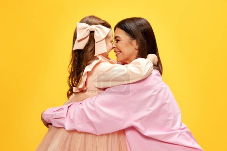 Photo for Portrait of beautiful young woman kissing her little pretty daughter, mother and kid posing in tender dresses against yellow studio background. Concept of happiness, Mothers day, childhood, fashion - Royalty Free Image