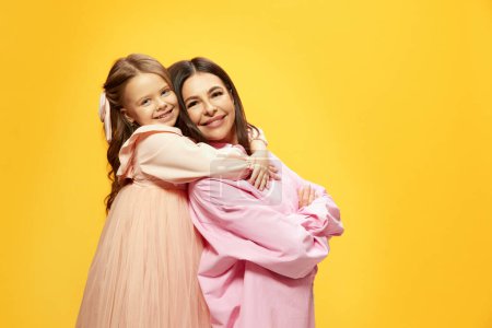 Photo for Happy, positive little girl ,daughter hugging her beautiful young mother against yellow studio background. Concept of happiness, Mothers day, childhood, fashion and lifestyle, birthday - Royalty Free Image