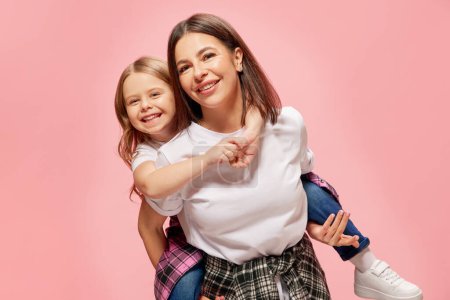 Photo for Joyful, happy, smiling mother giving her little daughter piggyback ride, playing against pink studio background. Concept of happiness, Mothers day, childhood, fashion and lifestyle - Royalty Free Image