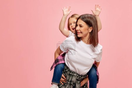 Foto de Alegre, feliz, sonriente madre dando a su pequeña hija paseo a cuestas, jugando contra el fondo del estudio rosa. Concepto de felicidad, Día de las Madres, infancia, moda y estilo de vida - Imagen libre de derechos