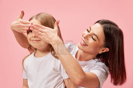Photo for Smiling beautiful young woman, mother covering her little daughters eyes, surprising and playing with kid against pink background. Concept of happiness, Mothers day, childhood, fashion and lifestyle - Royalty Free Image