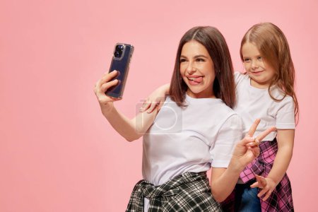 Photo for Positive, happy young mother takin selfie with her little adorable girl, daughter against pink studio background. Blog about motherhood. Concept of happiness, Mothers day, childhood, fashion - Royalty Free Image