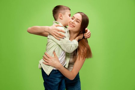 Photo for Happy, smiling little boy, son hugging and kissing his beautiful, caring mother, showing his love against green studio background. Concept of happiness, Mothers day, childhood, fashion and lifestyle - Royalty Free Image