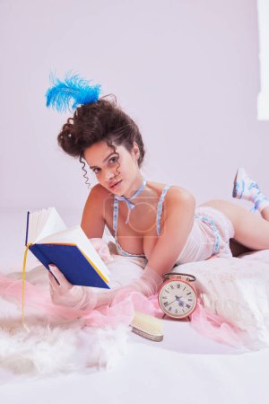 Photo for Attractive young woman in tender lingerie with feathered headpiece lying on bed with book. Vintage boudoir aesthetics. Concept of beauty and fashion, vintage, boudoir style - Royalty Free Image