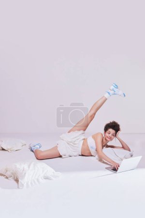 Téléchargez les photos : Élégante jeune femme couchée sur le lit avec ordinateur portable, jambe tendue, regarder la série télévisée sur fond pastel. Publicité pour défi de fitness virtuel. Concept de beauté et de mode, vintage, style boudoir - en image libre de droit