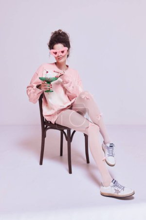 Téléchargez les photos : Jeune jolie femme assise sur une chaise en rose vêtements confortables et lunettes de soleil, manger du gâteau. Confort à la maison. Équilibre de vie. Concept de beauté et de mode, vintage, style boudoir - en image libre de droit