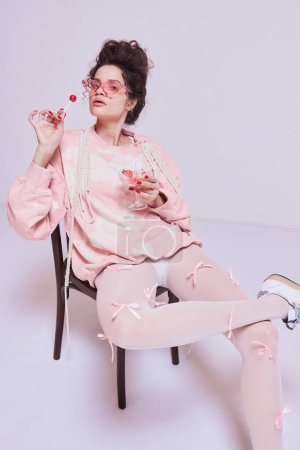 Téléchargez les photos : Élégante jeune femme élégante en tenue de maison rose confortable assise sur une chaise et mangeant des sucettes sur fond pastel. Concept de beauté et de mode, vintage, style boudoir - en image libre de droit