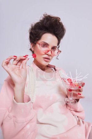 Foto de Elegante mujer joven con estilo en casa rosa cómoda llevar sentado en la silla y comer piruletas sobre fondo pastel. Anuncio de confitería. Concepto de belleza y moda, vintage, boudoir style - Imagen libre de derechos